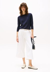 Jūras zils garām piedurknēm tops kopā ar augstas jostasvietas baltām plaša piegriezuma biksēm un bēšām loaferiem. Mazs melns pleca somiņa papildina apģērbu.