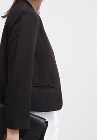 Femme en blazer sombre et pantalon blanc tenant une pochette en cuir noir sur un fond blanc uni.