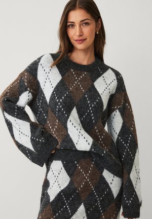 Kobieta ubrana w miękki sweter w argyle’owy wzór oraz dopasowaną spódnicę w kolorach ciemnoszarym, brązowym i białym, stojąca na tle jednolitego jasnego tła.