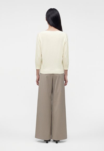 Femme aux longs cheveux noirs portant un haut crème à manches longues et un pantalon beige à jambes larges, debout de dos sur un fond uni.