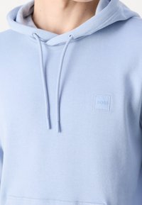 Světlé modrá mikina s kapucí a šňůrkami, s malou čtvercovou nášivkou s logem v levém horním rohu. Hladká textura látky.