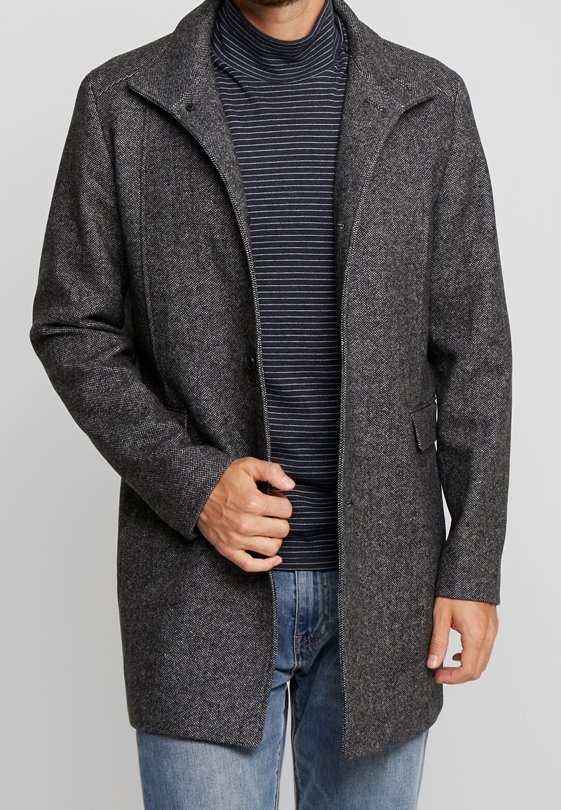 Abrigo gris de mezcla de lana con acabado texturizado, cierre de botones, cuello con muesca, bolsillos laterales y un ajuste ligeramente relajado, combinado con un suéter de cuello alto a rayas.