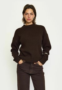 Brun stickad tröja med en ribbad textur, med rund hals och nedslagna axlar, kombinerad med mörkbruna byxor. Silverarmband som accent.