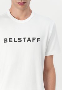 Fehér pamut póló, kerek nyakkivágással, fekete nyomott "BELSTAFF" felirattal a mellkasán. Puha tapintású, laza szabású.