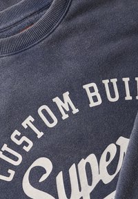 Sudadera de color azul marino con cuello de canalé; presenta texto impreso en blanco "CUSTOM BUILT" y "Super" en una tipografía en negrita. Textura suave.