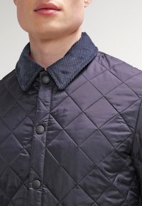 Nahaufnahme einer Person, die eine dunkle gesteppte Jacke mit einem Kordkragen und Druckknöpfen trägt.