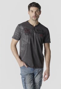 Homme aux cheveux courts et foncés, portant un polo à motif sombre et un jean cargo bleu clair, debout avec une main dans la poche sur fond blanc.