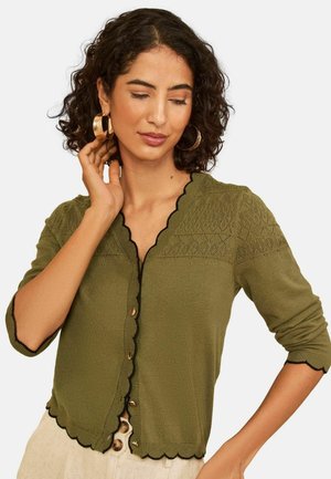 Frau mit lockigem dunklem Haar, die goldene Creolen und eine olivgrüne, geknöpfte Strickjacke mit wellenförmigen Kanten trägt.