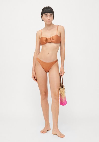 Brązowy bikini z przekręcanym przodem, cienkimi ramiączkami i dopasowanymi, niskimi majtkami. Modelka trzyma różową torbę ze słomy, boso.