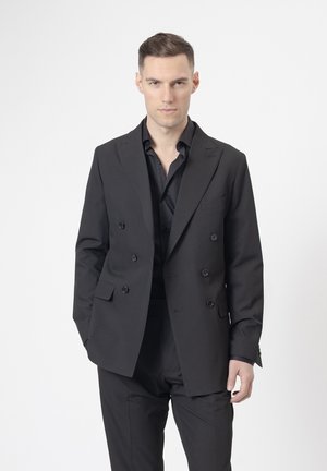 Hombre con cabello corto, vistiendo un blazer negro cruzado y una camisa negra, de pie frente a un fondo blanco liso.