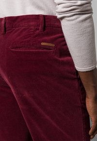 Burgundské manšestrové kalhoty s vertikálním žebrováním, s zadní kapsou a malým kovovým logem značky na pasu.