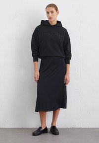 Felpa nera con logo "Marc O'Polo" in rilievo, abbinata a una gonna midi nera. Il modello indossa mocassini neri su uno sfondo neutro.