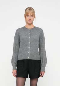 Cárdigan de lana gris con escote redondo, cierre de botones y mangas largas. Combinado con pantalones cortos negros con textura.