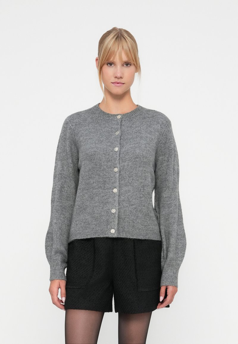 Cárdigan de lana gris con escote redondo, cierre de botones y mangas largas. Combinado con pantalones cortos negros con textura.