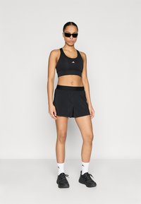 Sujetador deportivo negro con logo blanco, pantalones cortos negros a juego, calcetines blancos hasta el tobillo y zapatillas deportivas negras. Modelo con gafas de sol negras.