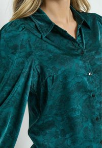 Türkise Button-up-Hemd aus Satin, mit floralem Muster, spitzem Kragen und schwarzen Knöpfen an der Knopfleiste. Puffärmel sichtbar.