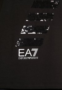 Černé bavlněné tričko s flitrovou grafikou v pruzích a bílým logem "EA7 EMPORIO ARMANI" ve spodní části. Hladká textura, jednoduchý design.