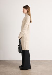 Beige színű, kötött pulóverruha magas gallérral, fekete, bő szárú nadrág fölött viselve. A modell egy fekete kézitáskát tart. Simább textúra.
