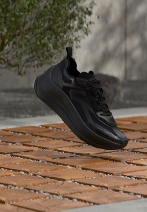 Zwarte Calvin Klein sneaker met dikke zool die zweeft boven houten tegels buiten, met groen gebladerte en een grijze muur op de achtergrond.