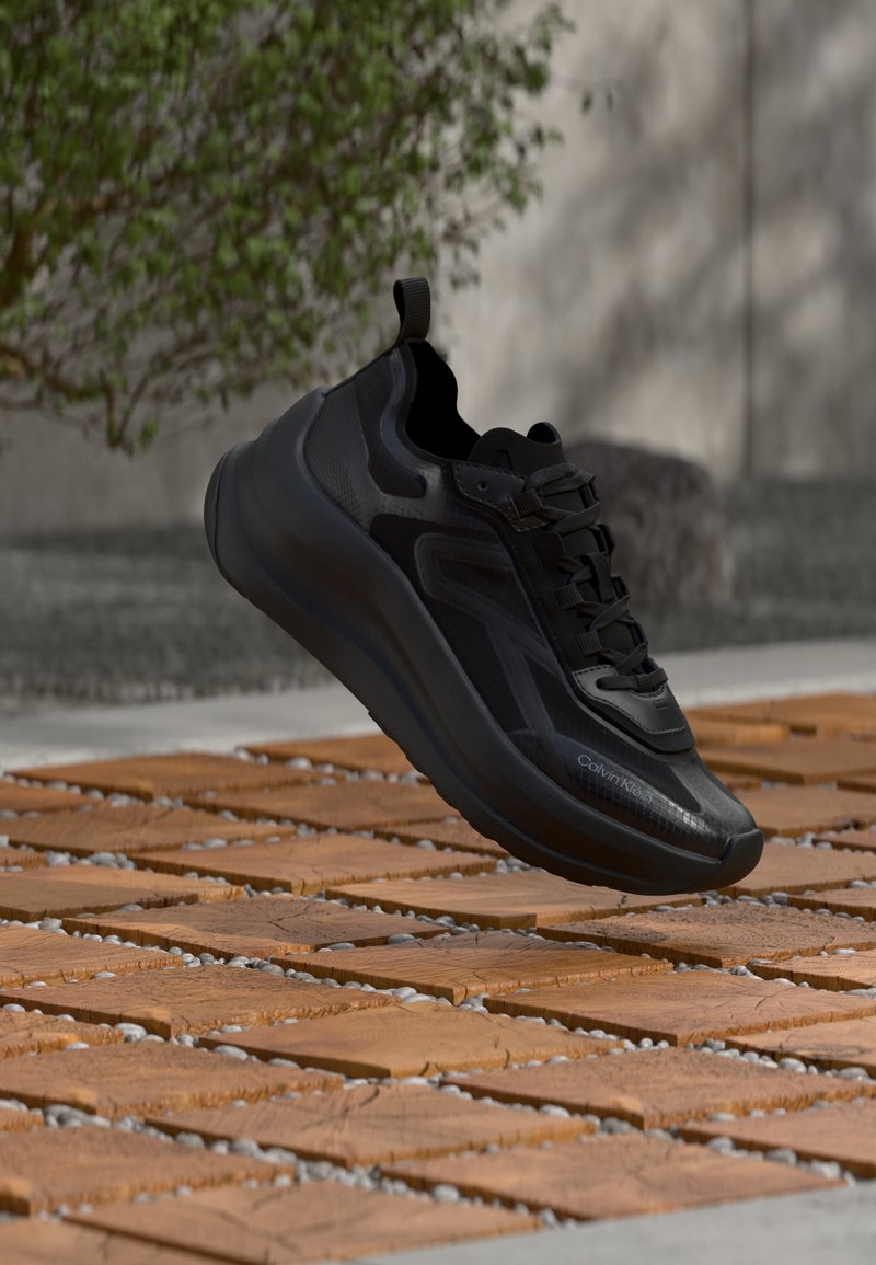 Baskets noires Calvin Klein à semelle épaisse flottant au-dessus de carreaux en bois en extérieur, avec un feuillage vert et un mur gris en arrière-plan.