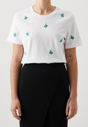 Femme portant un t-shirt blanc avec de petits cactus verts et des fleurs roses brodés, associé à une jupe noire taille haute.