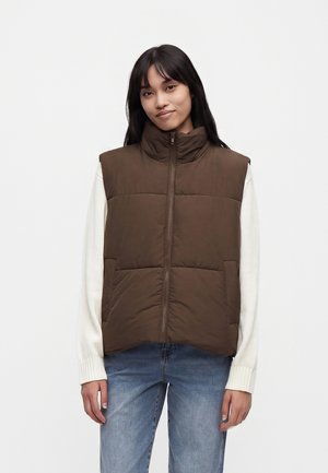 Junge Frau mit langen schwarzen Haaren, die eine braune Steppweste über einem weißen Pullover und blauen Jeans trägt und vor einem neutralen Hintergrund steht.