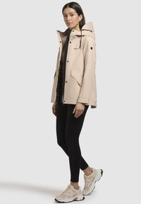 Beige waterdichte jas met een capuchon, voorzakken en verstelbare trekkoorden, gecombineerd met zwarte leggings en beige sneakers.