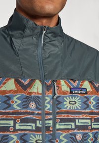 Patagonia jakke med en struktureret grå øverste del og en flerfarvet mønstret nederste del, med lynlåslukning og logo.