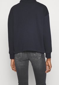 Felpa blu navy con maniche lunghe e orlo a coste, abbinata a jeans grigi scuri con cuciture a contrasto e tasche posteriori.