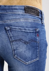Jeans de mezclilla azul que presentan un bolsillo trasero con costura en contraste y una etiqueta de cuero, mostrando una textura ligeramente desvaída.