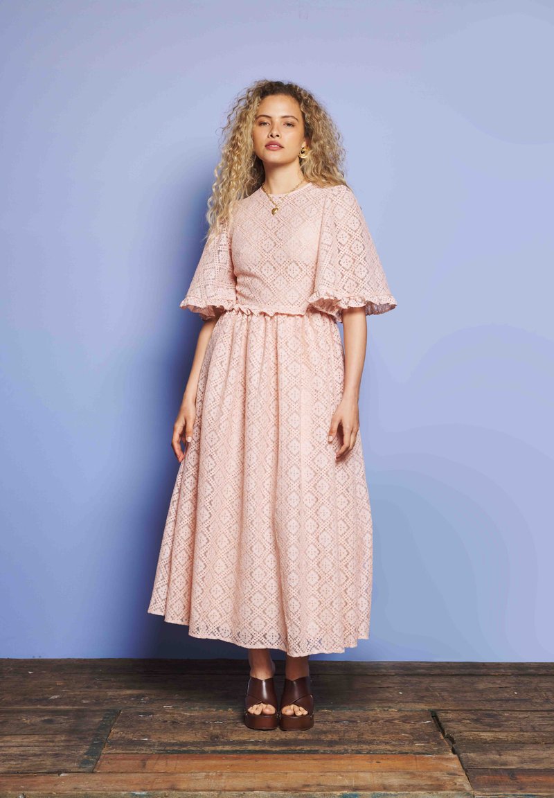 Mulher com cabelo encaracolado a pé em chão de madeira, vestindo um vestido midi estampado rosa com mangas largas e sandálias de plataforma castanhas.