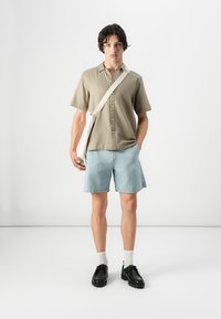 Chemise à manches courtes olive, short bleu clair, chaussettes blanches, chaussures noires et sac à bandoulière couleur crème. Design et coupe simples.