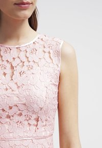 Růžové krajkové šaty s květinovým vzorem, bez rukávů, a hladkým saténovým výstřihem. Textura je jemná a komplikovaná.