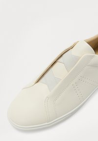 Białe skórzane buty wsuwane z elastycznymi krzyżującymi się paskami, perforowanymi detalami i zaokrąglonym noskiem. Gładka tekstura i minimalistyczny design.