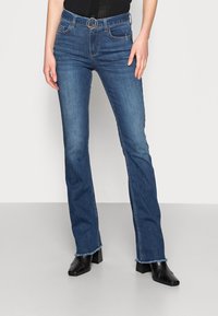 Kobieta w niebieskich dżinsach bootcut o średnim stanie z postrzępionymi brzegami, czarnym pasku, czarnej bluzce oraz czarnych botkach na obcasie z kwadratowym noskiem.