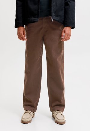 Homme portant un pantalon large marron, des chaussures décontractées beiges et une veste noire, debout devant un fond uni.