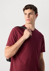 Jonge man in een bordeauxrood T-shirt die een zwart rugzakband over zijn schouder houdt, kijkend naar de zijkant tegen een effen achtergrond.