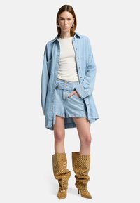 Lichtblauwe spijkerjurk over een witte top, gecombineerd met lichtblauwe shorts en bruine kniehoge laarzen met witte stippen.