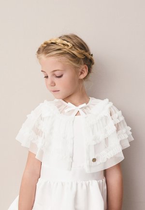 Blond dievča s pleteným venčekom, nosí biele šaty s volánikmi, pozerá dolu na jednoliatom béžovom pozadí.