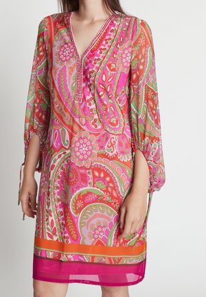 Woman wearing a knee-length dress with pink, orange, and green paisley patterns, sheer sleeves, and a V-neckline with tie details.
