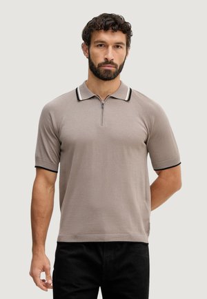 Partainen mies, joka on pukeutunut taupenväriseen, lyhythihaiseen neulepolo-paitaan, jossa on puolivetoketju ja mustat reunukset kauluksessa, seisoo yksinkertaista taustaa vasten.