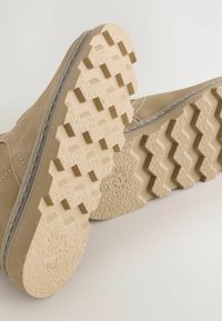 Beige mockaskinnsko med en texturerad gummisula som har zigzagmönster och en formad sulstruktur för grepp. Detaljerad söm längs ovansidan.