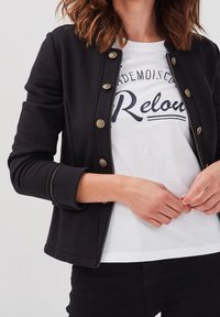 Woman wearing a black buttoned jacket over a white t-shirt with "Mademoiselle Relou" text, adjusting her hands near her waist.