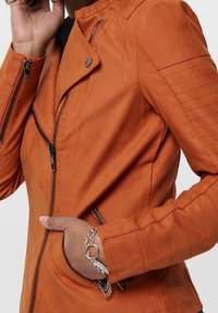 Pomarańczowa skórzana kurtka motocyklowa z zamkami błyskawicznymi, teksturowanymi szwami na ramionach oraz żebrowanymi detalami na rękawach. Akcenty z metalowych elementów.