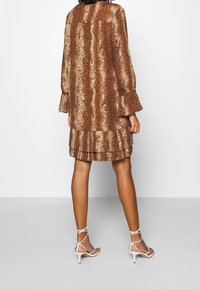 Braunes gemustertes Kleid mit einem eingefassten Saum, langen Ärmeln und einem lockeren Schnitt. Kombiniert mit weißen Riemchen-Heels.