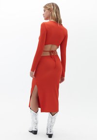 Rotes geripptes Zweiteiler-Set mit einem cropped Oberteil mit offener Rückseite und Bindungsdetail, kombiniert mit einem Rock mit hohem Schlitz. Weiße verzierte Stiefel.