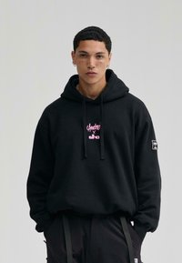 Sudadera con capucha negra con un logo gráfico rosa en el pecho. Cuenta con cordones y un logo de parche en la manga. La tela parece suave.