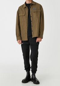 Camicia verde oliva con bottoni, dotata di due tasche sul petto e una toppa nera, abbinata a pantaloni cargo neri e stivali neri chunky.