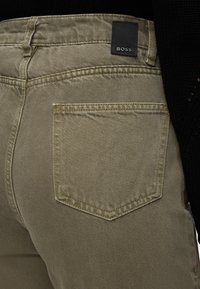 Gros plan sur la poche arrière d'un pantalon beige avec une étiquette noire de la marque Boss à la taille, porté avec un top en maille noire.