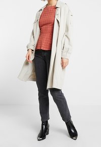 Trench-coat beige porté sur un haut perforé couleur rouille, associé à un jean gris foncé et des bottines noires à bouts pointus.